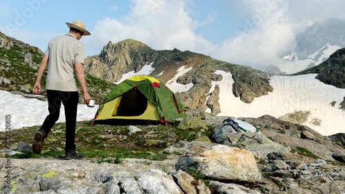 Solo Camper Walking Toward Green Tent on Rocky Mountain Snowfield in Remote Alpine Landscape