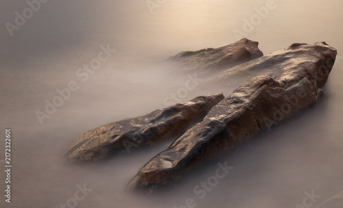 Long-exposure detail of smooth coastal rocks emerging from silky Mediterranean water on the rugged shoreline of Porto, Corsica, France.