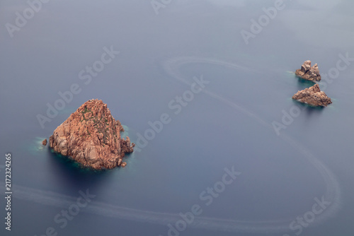 Three red granite sea stacks rising from calm Mediterranean water off the rugged western coast of Corsica, France, photographed with a long exposure.