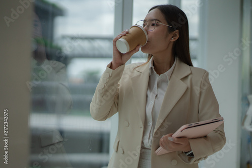 Foto Business lifestyle concept of a smiling young woman holding coffee and tablet by an office window during a work break