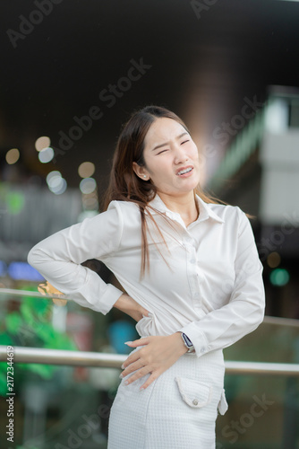Fotografie Back pain concept of a young businesswoman holding her lower back with discomfort at work, showing muscle strain and fatigue