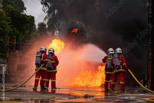 Canvas Print Professional firefighters in full protective gear bravely battle a massive, blazing inferno, holding a hose as intense orange flames and smoke consume the background in an emergency
