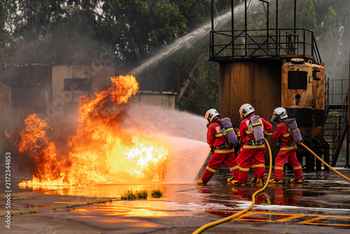 Canvas Print Professional firefighters in full protective gear bravely battle a massive, blazing inferno, holding a hose as intense orange flames and smoke consume the background in an emergency