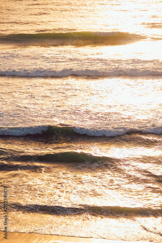 Waves are rolling gently on the beach as the sun sets. The sky reflects warm colors on the water and the scene shows a calm coastal view with distant hills.