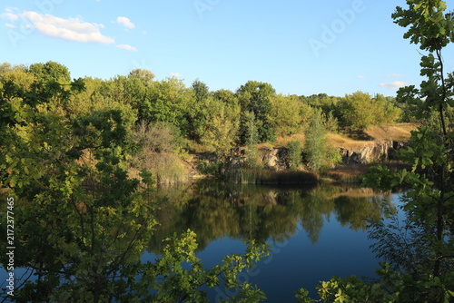 Scenic landscape of a rocky quarry lake with clear water, surrounded by trees and dry grass on a sunny summer day