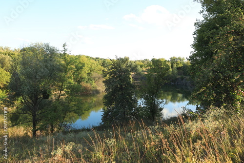 Scenic landscape of a rocky quarry lake with clear water, surrounded by trees and dry grass on a sunny summer day