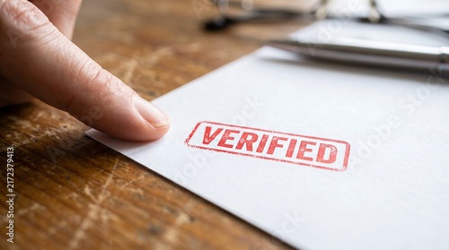 Closeup of a finger pointing at a red verified stamp on a white paper document on a wooden desk