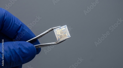 Closeup of a gloved hand holding a small computer chip with tweezers against a grey background