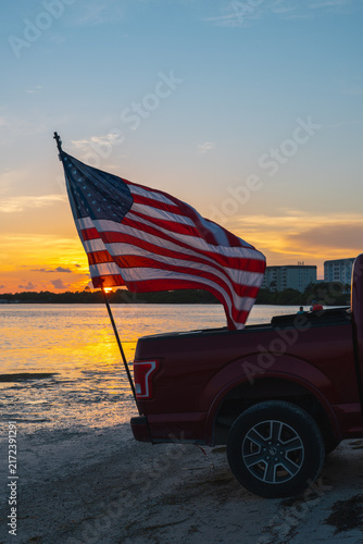 American flag. United States flag waving from a pickup truck by the water during a colorful sunset. Patriotic USA symbol with red, white and blue stars and stripes. American lifestyle. State Florida