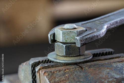 Macro of open end wrench turning rusty steel nut on bolt with washer clamped in heavy metal vice. Mechanics work, workshop repair, industrial tools and hardware concept