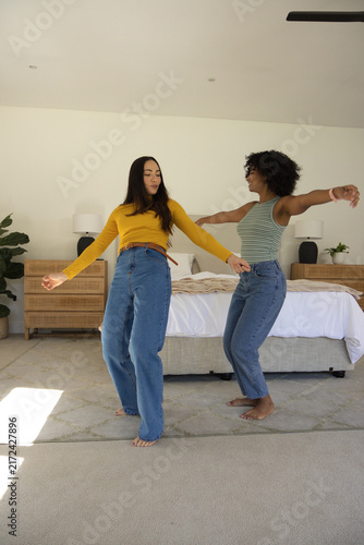 Diverse female friends dancing in bright bedroom on area rug by bed, wearing jeans, barefoot