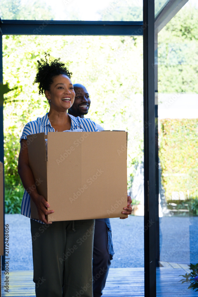 Naklejka premium African American couple carrying large plain cardboard box through glass doorway onto wooden deck