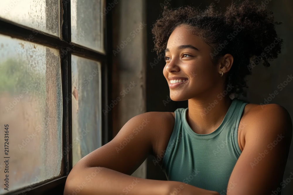Fototapeta premium Smiling young woman looking out of a window with natural sunlight illuminating her happy face