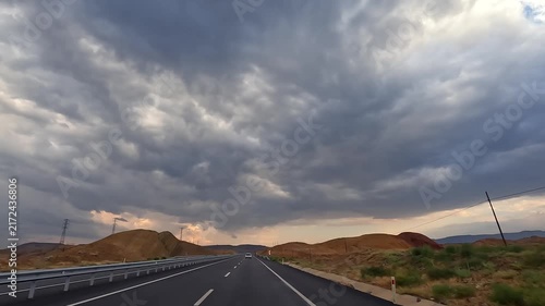 Car traveling along highway, wide view, through open countryside in Turkey beneath storm clouds, showing long-distance mobility, changing weather, road infrastructure, self-drive tourism