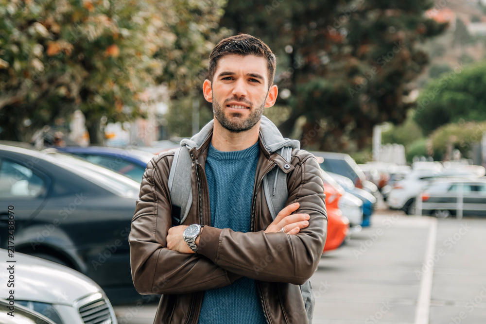 Obraz premium Confident young man with crossed arms standing outdoors in a parking lot on a sunny autumn day, smiling with a casual travel and tourism lifestyle concept