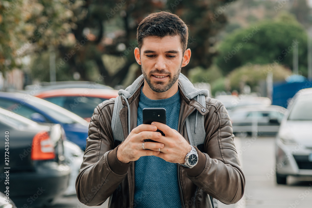 Obraz premium Young man with beard smiling while checking smartphone on urban street with parked cars in background