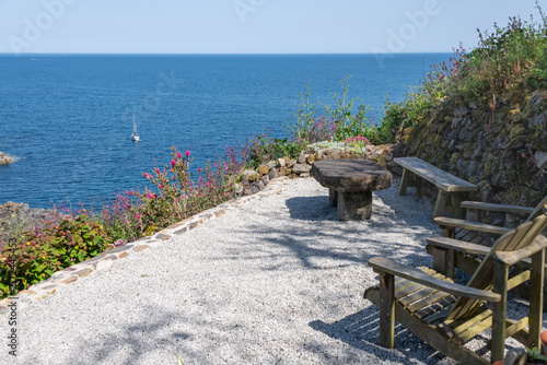 Seating in a coastal garden, overlooking the sea, cliffs, and blue sky. With summer flowers and a yacht in the background.
