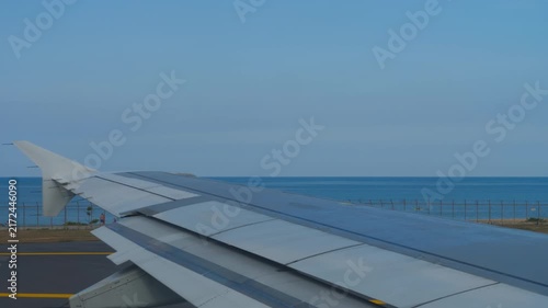 Airplane wing moving on airport runway next to blue ocean and Mai Khao Beach under clear blue sky daytime shot