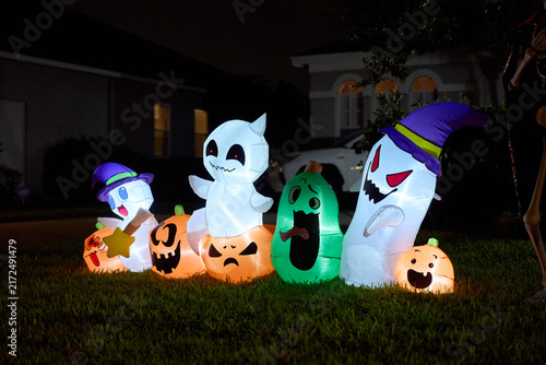 A lineup of smiling ghosts and carved pumpkin inflatables lights a suburban yard. Festive Halloween scene with vivid colors after dark.