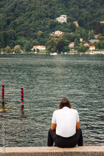 Un homme assis de dos face à un lac. Homme réfléchissant face au Lac Majeur. Solitude en vacances. Nostalgie et tristesse humaine