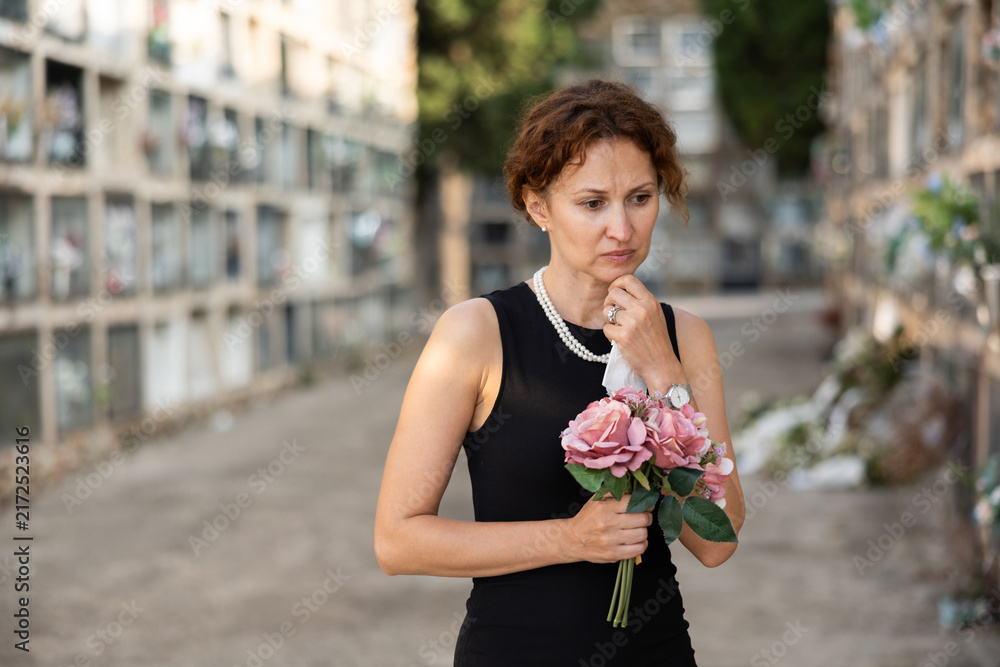 Fototapeta premium Sorrowful middle aged woman in black dress standing in cemetery holding bouquet of flowers with reflective and somber expression against backdrop of stone memorial walls