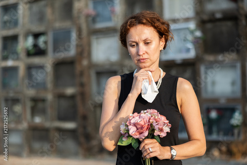 фотографія Sad woman in black mourning dress and pearl necklace standing in front of columbarium in old graveyard holding fresh flower bouquet, immersed in grief