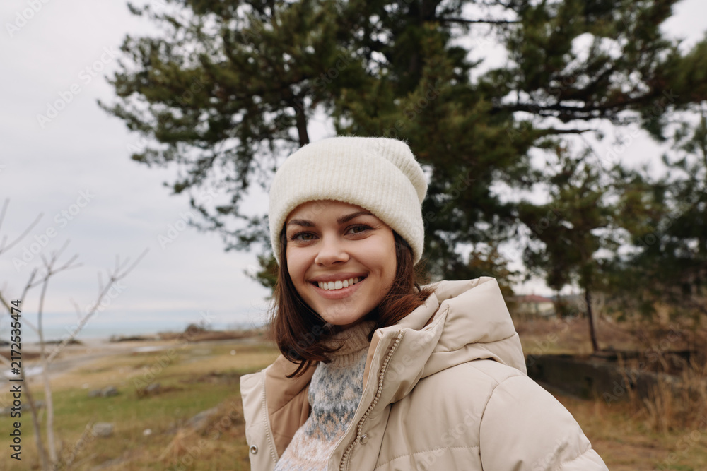 Fototapeta premium Woman in a cozy beige jacket and knit beanie outdoors, smiling with a scenic field and distant water behind her, amid trees and crisp air.