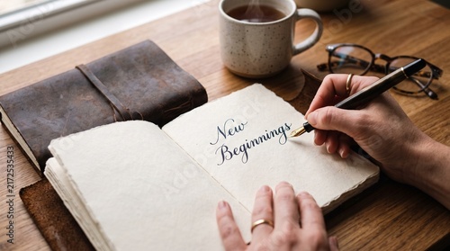 Hands writing 'New Beginnings' in a journal beside a coffee cup and leather-bound notebook.