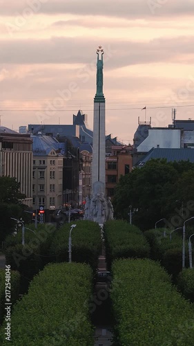 An aerial view showcases the Freedom Monument rising above a tree lined boulevard in Riga, Latvia during a peaceful sunset.