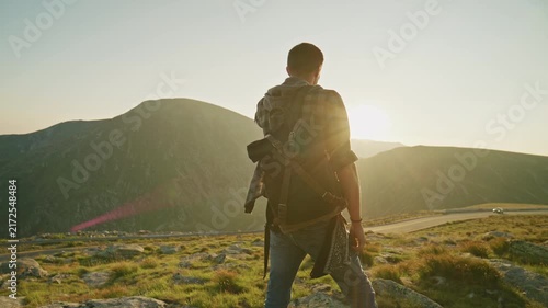 Hiker stands on a mountain looking at the sunset over hills and a road in the distance