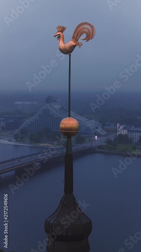 Aerial view showing the golden rooster weather vane of Riga Cathedral spire with the Daugava River and cityscape at dusk, Latvia.