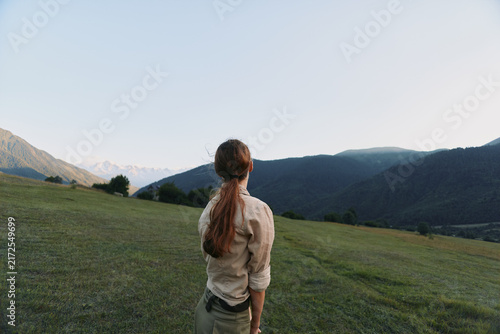 Young woman with long hair stands on green hilltop looking at mountain range under clear sky, enjoying peaceful nature and outdoor lifestyle moment.