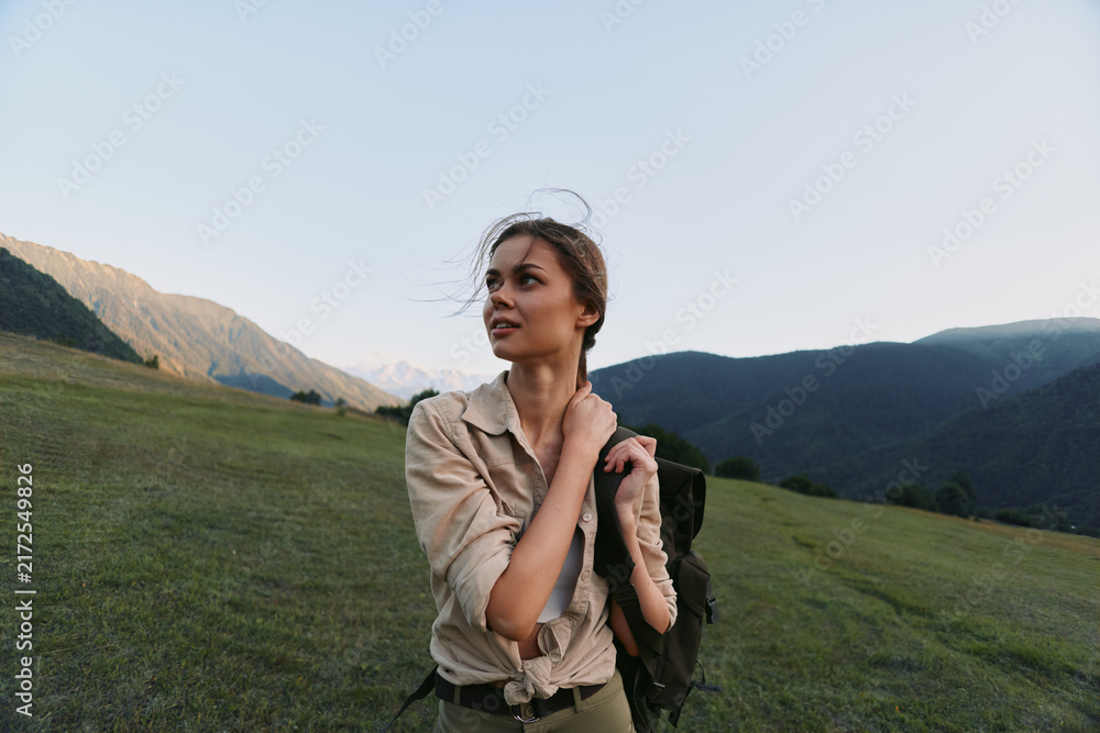 Fototapeta premium Young woman traveler stands on green mountain meadow with backpack looks away thoughtful enjoying nature and fresh air during hiking trip in clear sky daylight.