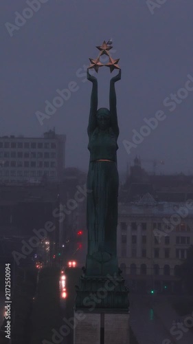 An aerial perspective captures the Freedom Monument in Riga, Latvia, standing above urban street traffic during foggy twilight conditions.