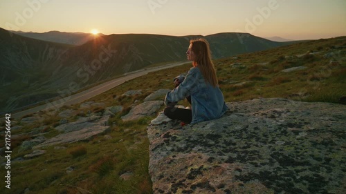 Sunset view from a rocky ledge with a woman sitting on the ground while looking at the horizon in a mountain area
