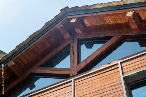 Construction detail: wood truss, gable roof, angled view from below, beams and pillars, dark wood, laminated wood, cladding panels, horizontal, curtain wall, glass, blue sky