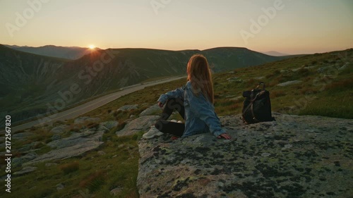 Woman sits on a rock looking at sunset over mountains and road in the evening