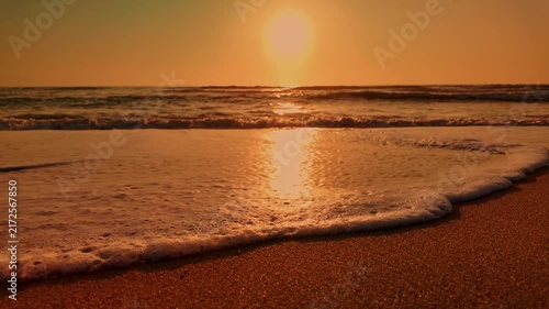 A low angle view captures gentle ocean waves washing onto a wet sandy beach under a warm sunset, creating a tranquil and golden atmosphere with a glossy texture.