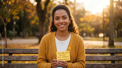 Mulher jovem sorridente sentada em banco de parque ao pôr do sol segurando cartão amarelo com mensagem motivacional, campanha Setembro Amarelo, saúde mental e esperança
