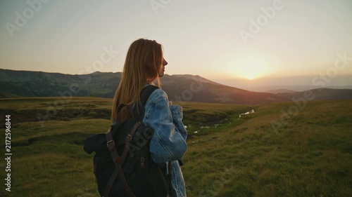 Woman stands on green hillside and watches sunset in mountainous area