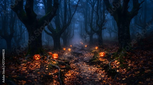 A mysterious spooky forest path at night illuminated by glowing carved Halloween pumpkins. Gnarled dark trees rise through thick blue fog and mist, with the forest floor covered in orange autumn fa...