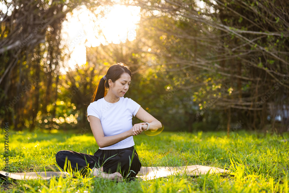 Obraz premium Woman sitting on a yoga mat looking down at her smartwatch in a sunny park