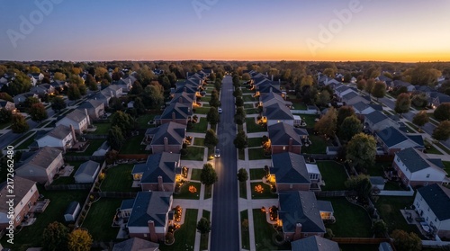 Aerial view of a suburban neighborhood at dusk with houses, illuminated windows, and streetlights along tree-lined streets under a warm sky.