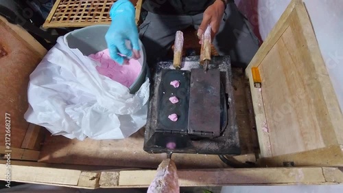 Person wears gloves, works with pink wax. Hands press wax into wooden mold. Wax melts in heated, rustic container. Gloved hands guide the process carefully. Traditional craft captured in close-up view
