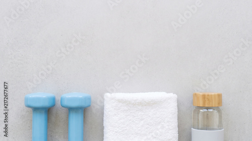 Minimalist fitness composition featuring light blue dumbbells white gym towel and glass water bottle on a clean grey concrete background for healthy lifestyle and wellness branding concepts