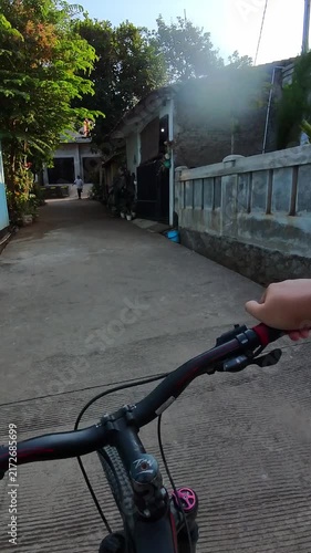 Vertical POV video of Bicyclist rides through quiet village streets. Sunlight casts warm glow on narrow roads. Simple houses line the path with greenery. Handlebars frame view as cyclist moves forward