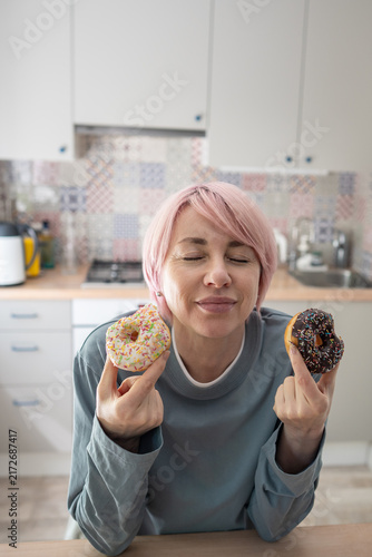 Foto Mature woman holds two colorful donuts with closed eyes, surrendering to intense PMS cravings and sugar addiction after trying to avoid sweet pastries and maintain dietary self-control