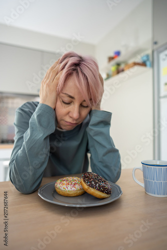 Obraz na plátne Mature woman sits distressed before colorful donuts, struggling with PMS, sugar addiction and pastry cravings while trying to avoid sweets and maintain self-control during difficult withdrawal