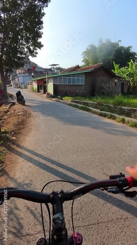 Vertical Video of Bicyclist rides down quiet residential street. Handlebars frame view of narrow road ahead. Trees cast long shadows on paved surfaces. Hand grips bike as it moves forward gently. 4K 
