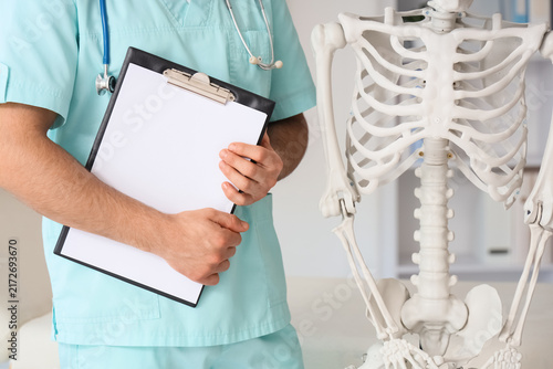 Foto Male doctor with clipboard and human skeleton in clinic, closeup
